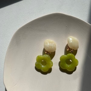 Op de afbeelding: Een paar groene en witte bloemvormige oorbellen met gouden clip-sluitingen. De oorbellen liggen op een wit oppervlak.