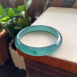 Puede incluir: Un brazalete tipo esclava liso y translúcido en un tono verde azulado vibrante. El accesorio circular presenta una superficie pulida y redondeada con variaciones sutiles en la profundidad y claridad del color, mostrando un diseño de banda sólida.