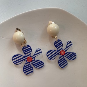 Peut inclure: Une paire de boucles d'oreilles en forme de fleur à rayures bleues et blanches avec un centre rouge. Les boucles d'oreilles sont attachées à une perle blanche.