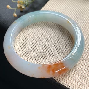 Könnte beinhalten: Ein glatter, kreisförmiger Armreif aus durchscheinendem Stein. Das Schmuckstück hat eine blass, eisig blau-grüne Grundfarbe mit einem markanten, leuchtend orange-roten Farbfleck, der sich auf einer Seite des polierten, abgerundeten Bandes befindet.