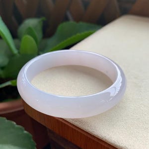 May include: A smooth, translucent, pale lavender-white bangle bracelet with a rounded profile. The circular jewelry piece features a polished, glossy surface that reflects light, showcasing a consistent, solid color throughout its entire structure.