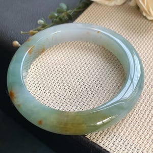 Könnte beinhalten: Ein glatter, polierter Jade-Armreif mit einer durchscheinenden hellgrünen Basis, subtilen wolkigen weißen Einschlüssen und markanten, kleinen bernsteinfarbenen Mineralflecken, die über die abgerundete Oberfläche des kreisförmigen Schmuckstücks verteilt sind.