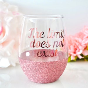 May include: A clear glass wine glass with a pink glitter rim. The glass has the text "The limit does not exist" printed on it in rose gold.