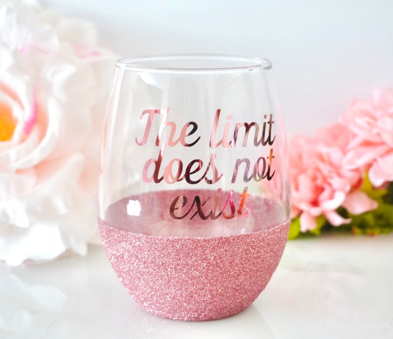 May include: A clear glass wine glass with a pink glitter rim. The glass has the text "The limit does not exist" printed on it in rose gold.