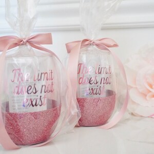 May include: Two clear glass wine glasses with pink glitter at the bottom. The glasses are wrapped in clear cellophane and tied with pink ribbon. The glasses have the text "The limit does not exist" printed on them.
