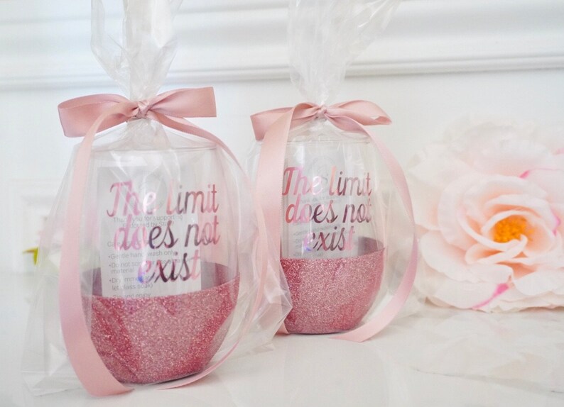 May include: Two clear glass wine glasses with pink glitter at the bottom. The glasses are wrapped in clear cellophane and tied with pink ribbon. The glasses have the text "The limit does not exist" printed on them.