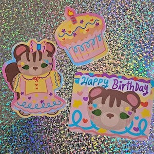 May include: Three colorful stickers with a birthday theme. One sticker features a cupcake with a lit candle. Another shows a bear character in a dress. The third sticker has the text "Happy Birthday" with a bear face.