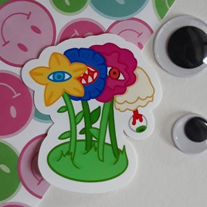 May include: A sticker featuring a whimsical illustration of several colorful flowers with eyes and teeth. The flowers are yellow, blue, and pink, with green stems and a green base. The background includes smiley faces and googly eyes.