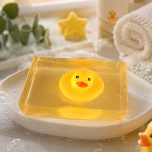 May include: A rectangular bar of translucent yellow soap featuring a small, embedded yellow rubber duck toy in the center. The soap has a smooth, clear texture and is resting on a white ceramic dish, showcasing a bright and cheerful appearance.