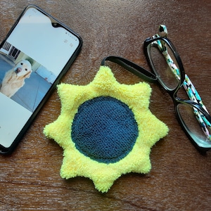 May include: A yellow and blue microfiber cleaning cloth shaped like a sunflower. The cloth is on a wooden surface with a pair of reading glasses and a smartphone.