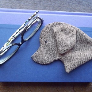 May include: A pair of black reading glasses with a floral pattern on the arms, lying on a blue and purple book. A brown fabric dog head is on the book.