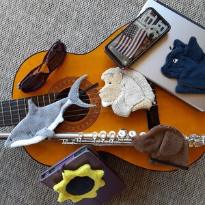 This is a group photo of different Smudge products that are available:  a shark, a lion, sunflower, black cat and chocolate lab. They are arranged with different items that they can be used to clean and polish: a guitar, a flute, tablet, etc.