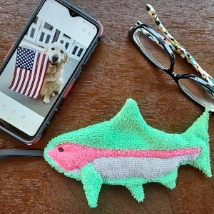 May include: A green and pink shark-shaped bath sponge with a white stripe. A smartphone with a photo of a golden retriever puppy holding an American flag is in the background.