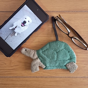 May include: A green and beige turtle-shaped washcloth with a black elastic loop, beside a smartphone showing a photo of a white dog and a pair of spectacles with a brown frame. The items are on a wooden surface.