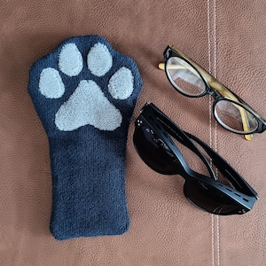 May include: A dark blue, paw-shaped, soft, textured hand warmer with a white paw print on the front. Two pairs of glasses are also in the image, one pair is black and the other is tortoise shell.