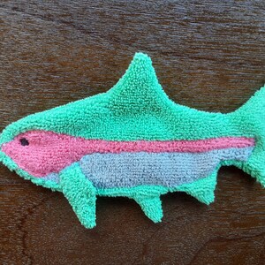 Peut inclure: Un jouet en peluche en forme de poisson vert, rose et gris. Le poisson a une nageoire sur le dos et une nageoire caudale. Le poisson a une ficelle noire attach&eacute;e &agrave; sa t&ecirc;te.
