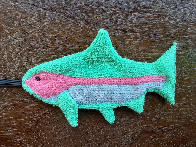 Peut inclure: Un jouet en peluche en forme de poisson vert, rose et gris. Le poisson a une nageoire sur le dos et une nageoire caudale. Le poisson a une ficelle noire attach&eacute;e &agrave; sa t&ecirc;te.