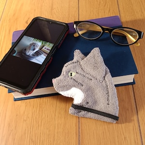 May include: A gray cat-shaped hand towel with a black elastic band. The towel is on a blue book with a black smartphone on top. The smartphone has a photo of a golden retriever puppy on the screen.