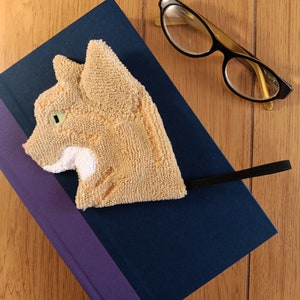 May include: A plush orange cat head keyring with a black strap. The keyring is lying on a blue and purple book with a wooden background.