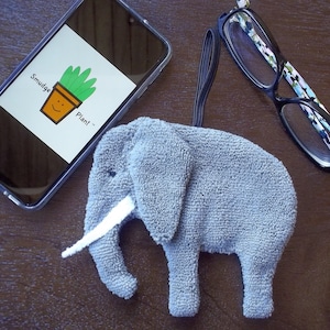 May include: A grey elephant-shaped microfiber cloth with a white tusk. The cloth is used for cleaning glasses. The text "Smudge Plant" is visible on a smartphone in the background.