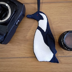 May include: A navy blue and white penguin-shaped ornament with a black strap. A black camera and a Canon lens are also on a wooden surface. The penguin has red accents and is made of a textured fabric.