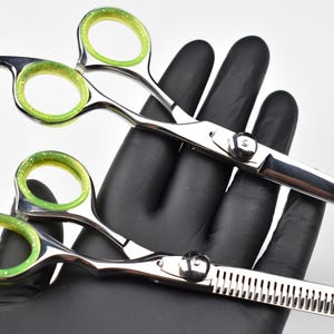 May include: Two professional hair shears with silver blades and handles. The finger holes are green with glitter. One shear has a serrated edge. The shears are held by a black gloved hand.