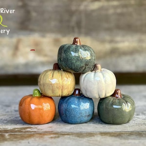 May include: A stack of six small ceramic pumpkins in various colors including orange, yellow, white, blue, and shades of green. Each pumpkin features a smooth, glossy glaze and a small brown stem. The text Leaf River Pottery is in the top left corner.