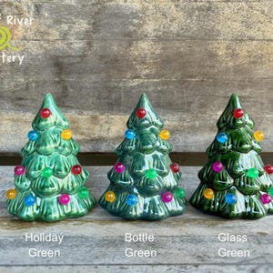 May include: Three ceramic Christmas trees in varying shades of green, each adorned with colorful, round lights. The trees are labeled "Holiday Green," "Bottle Green," and "Glass Green." The Leaf River Pottery logo is in the upper left corner.