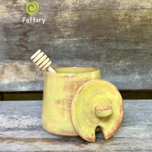 May include: A yellow ceramic honey pot with a matching lid featuring a small handle and a notch for a wooden honey dipper. The pot has a glazed finish with subtle brown undertones. The text Leaf River Pottery is visible in the upper left corner.
