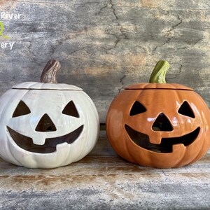 May include: Two ceramic jack-o'-lantern candle holders. One is white with a brown stem and the other is orange with a green stem.
