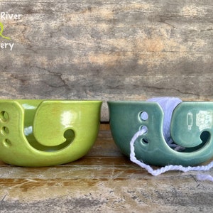 May include: Two ceramic yarn bowls in shades of green. The bowl on the left is a bright lime green, and the one on the right is a teal green. Both have a curved opening for yarn and holes for the yarn to pass through. The text "Leaf River Pottery" is in the upper left.
