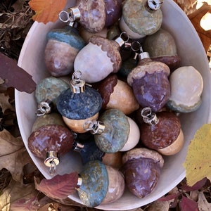 May include: A collection of ceramic acorn ornaments in a white bowl, set against a backdrop of autumn leaves. The acorns feature a variety of colors, including brown, blue, and purple, with silver metal hangers for easy display. The ornaments are handmade.