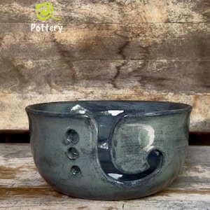May include: A dark blue-grey ceramic yarn bowl with a glossy finish. The bowl features a spiral cutout on the front for yarn and three small circular holes on the side. The text Leaf River Pottery is printed in white above the bowl.