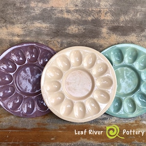 May include: Three ceramic egg dishes, one purple, one beige, and one teal green. Each dish has 12 indentations for eggs. The dishes are arranged in a semi-circle on a wooden surface. The text "Leaf River Pottery" is visible in the bottom center of the image.