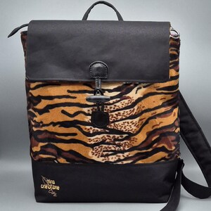Könnte beinhalten: Ein schwarzer Rucksack mit Tigerprint, Klappe und Griff. Die Tasche hat einen Knebelverschluss und verstellbare Träger. Der Boden der Tasche ist schwarz, und der Text "mins one of one" ist sichtbar.