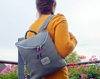 Könnte beinhalten: Ein grauer Canvas-Rucksack mit Reißverschluss und einem kleinen rechteckigen Etikett mit der Aufschrift "mini monochrome".