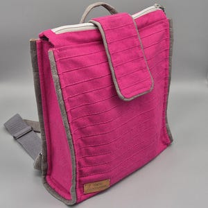 May include: A pink canvas backpack with a grey zipper and trim. The backpack has a small pocket on the front with a flap closure. The backpack has a label that says "mini kangaroo".