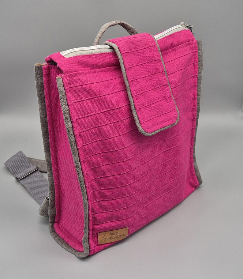 May include: A pink canvas backpack with a grey zipper and trim. The backpack has a small pocket on the front with a flap closure. The backpack has a label that says "mini kangaroo".