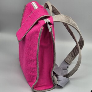 May include: A pink canvas backpack with grey straps. The backpack has a zipper closure and a small pocket on the front.