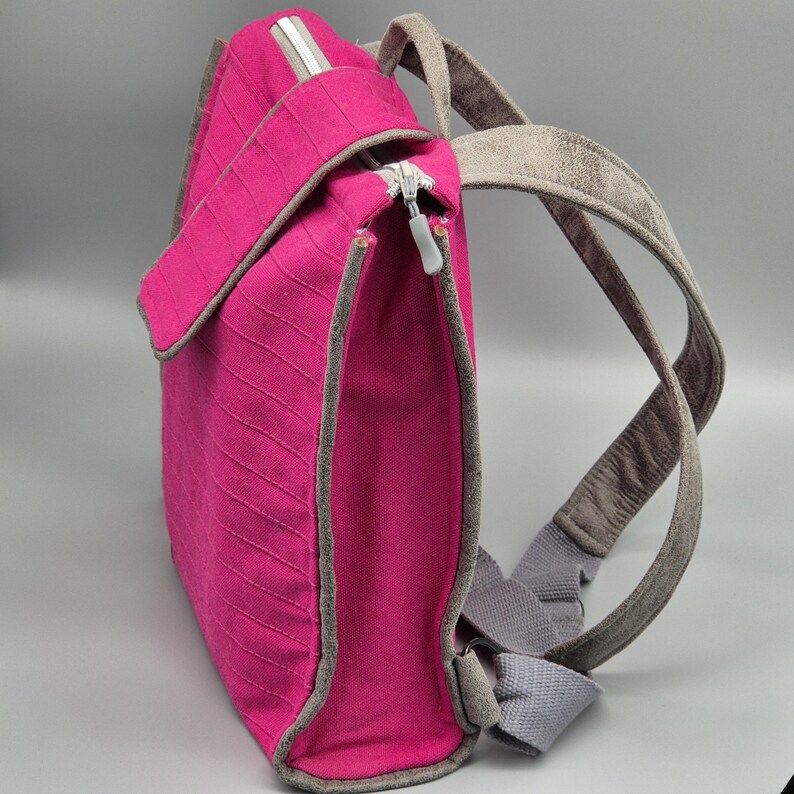 May include: A pink canvas backpack with grey straps. The backpack has a zipper closure and a small pocket on the front.