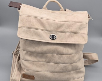 Könnte beinhalten: Ein beigefarbener Rucksack aus Wildleder mit einer Klappe und einem silbernen Knopf. Der Rucksack hat einen braunen Lederriemen und ein kleines braunes Lederetikett mit dem Text "INTO THE WILD" darauf.