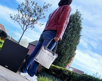 May include: A quilted off-white tote bag with grey trim and handles is held by a person wearing a red leather jacket and blue jeans. The bag has a zipped pocket and a small metal ring. The background includes a blue sky and greenery.