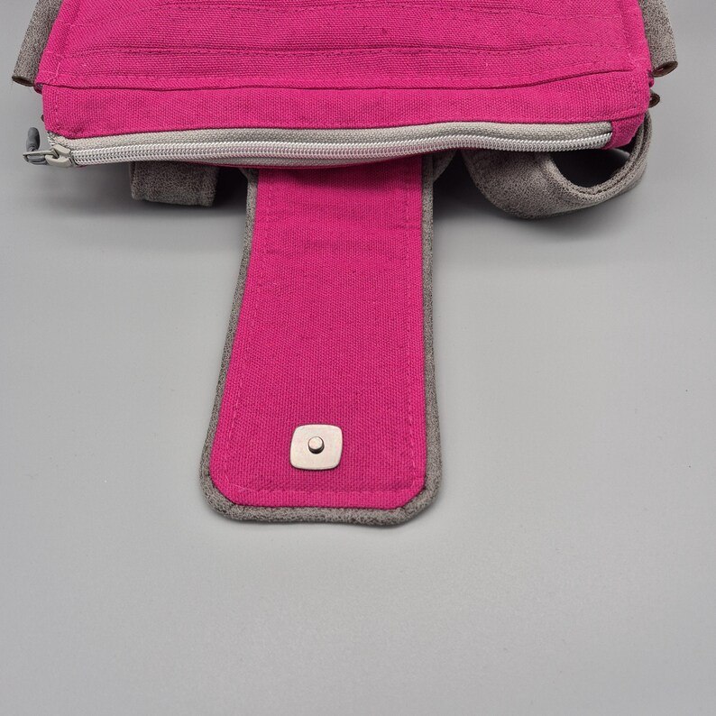 May include: A pink fabric bag with a zipper closure and a grey fabric strap. The strap has a metal button.
