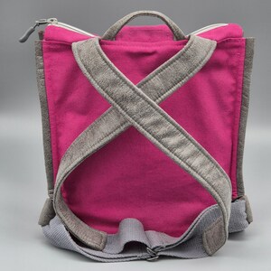May include: A pink canvas backpack with grey suede straps. The straps cross at the back and are adjustable.