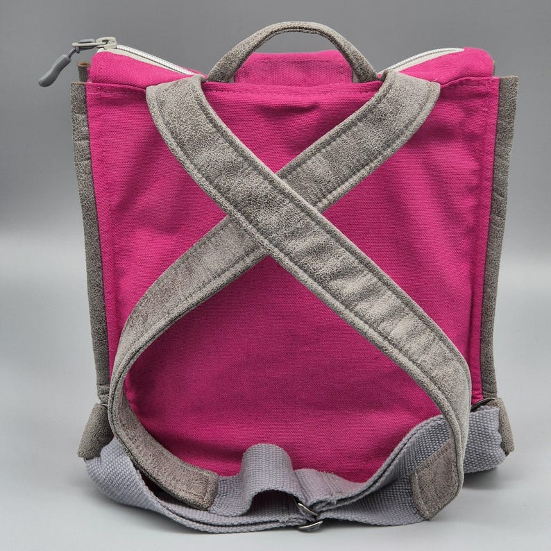 May include: A pink canvas backpack with grey suede straps. The straps cross at the back and are adjustable.