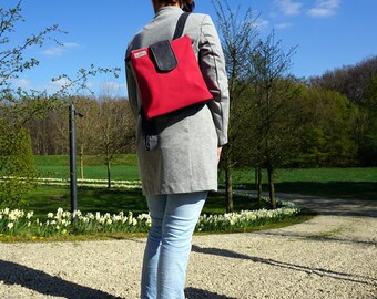 Könnte beinhalten: Eine Frau in einer grauen Jacke und blauen Jeans geht in einem Park spazieren. Sie trägt einen roten Rucksack mit einer schwarzen Klappe.