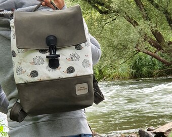 Könnte beinhalten: Ein grauer und weißer Rucksack mit einer Klappenverschlusses und einem Igel-Druck. Der Rucksack hat ein kleines Leder-Etikett mit dem Text "mama fontoura" darauf.