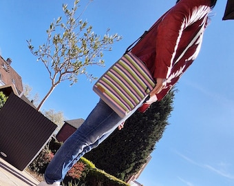 May include: A striped shoulder bag in shades of green, pink, and purple. The bag is being carried by a person wearing a red jacket and blue jeans. The background shows a clear blue sky and a small tree.