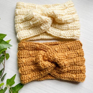 May include: Two knitted headbands, one in a cream color and one in a mustard yellow color. Both headbands have a knotted design.
