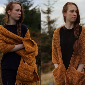 May include: Mustard yellow crocheted shawl with two pockets and fringe. The shawl has a textured stitch pattern and is draped over the shoulders. The pockets are also crocheted and have a similar texture. The fringe hangs from the bottom edge.